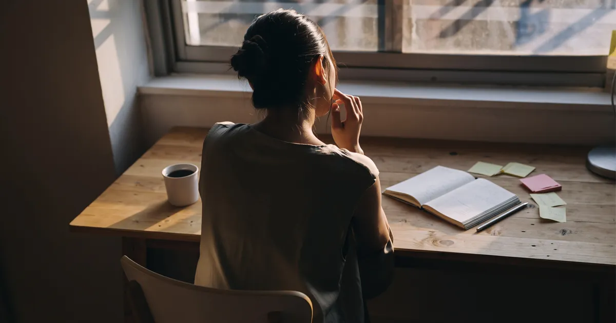 窓辺の机に座り、ノートと付箋を前に考え込む人物の後ろ姿