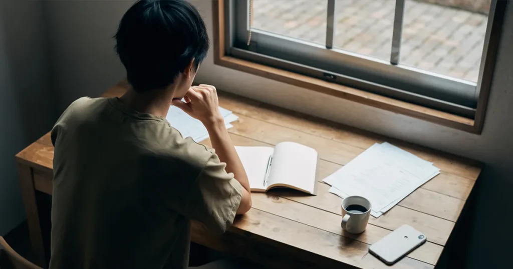 窓辺の机でスマホとノートを前に静かに座る人物の後ろ姿